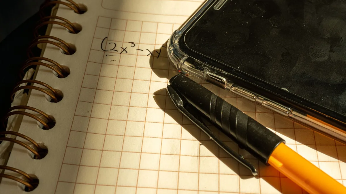 A notebook with a math equation, smartphone, and pen on checkered paper in warm lighting.