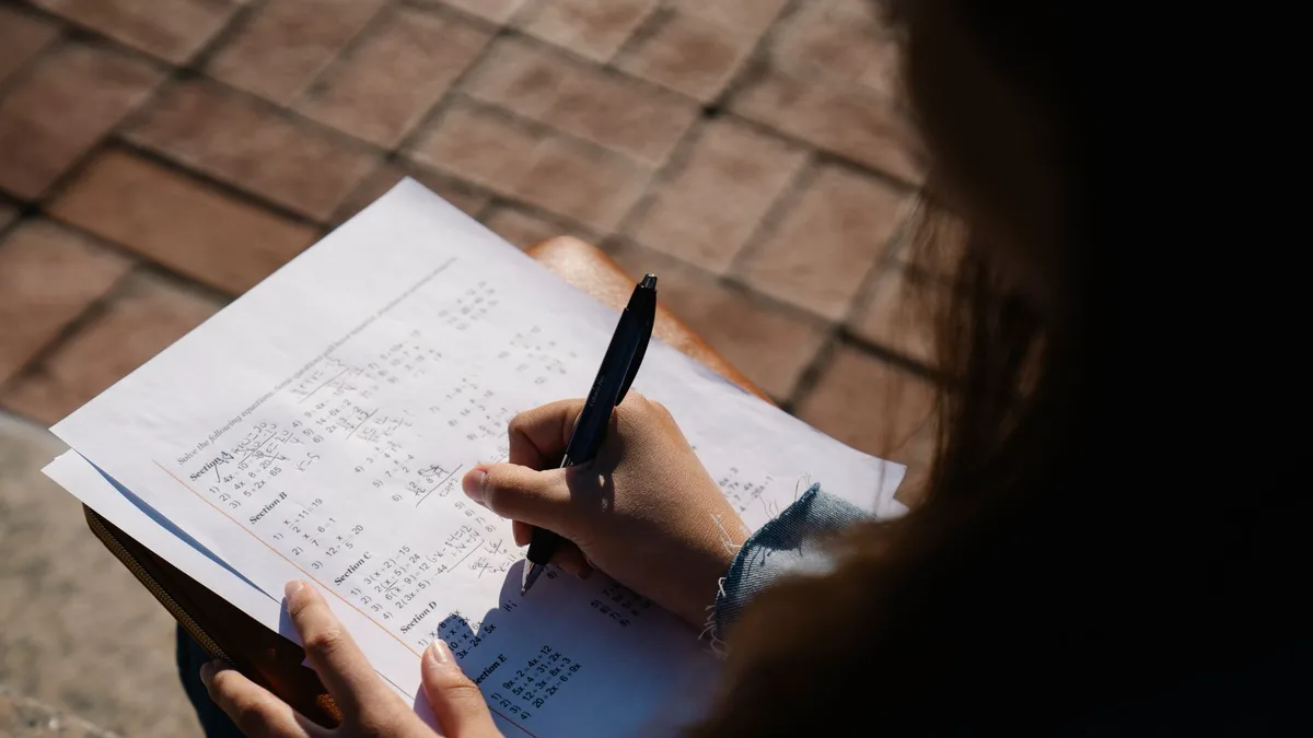 A person writing calculations on a mathematical worksheet with a pen outdoors.