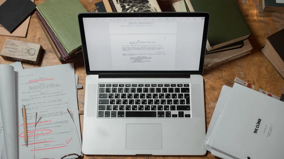 A laptop surrounded by books and scripts on a wooden desk, perfect for literature and technology themes.