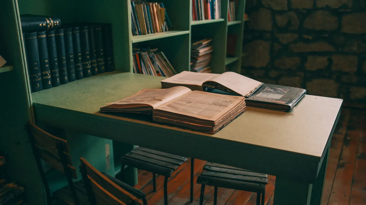 Warm-toned library interior featuring open books on a vintage table with green shelves.