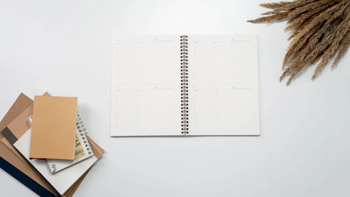 Open notebook with planner pages, surrounded by notebooks and pampas grass on a white background.